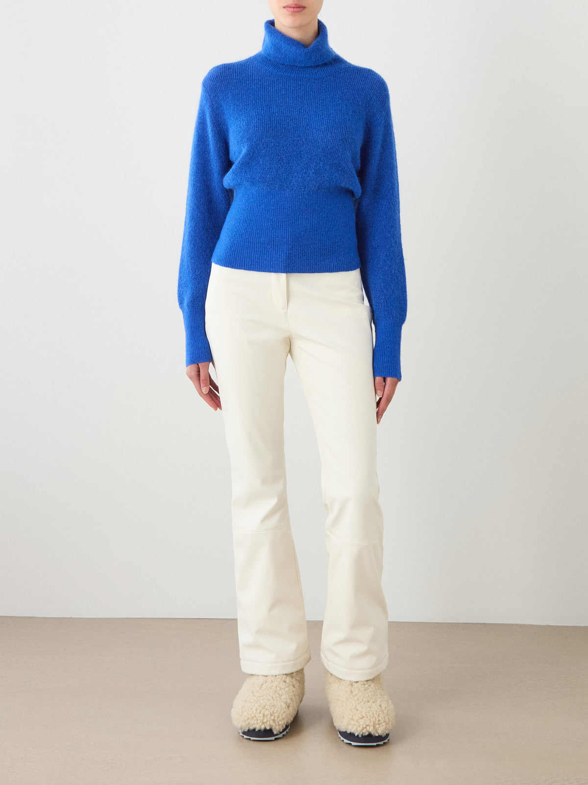 A person stands against a plain light-colored background wearing the We Norwegians Pudder Sweater Women—a bright blue turtleneck crafted from mohair yarn—paired with off-white pants and fuzzy beige slip-on shoes.