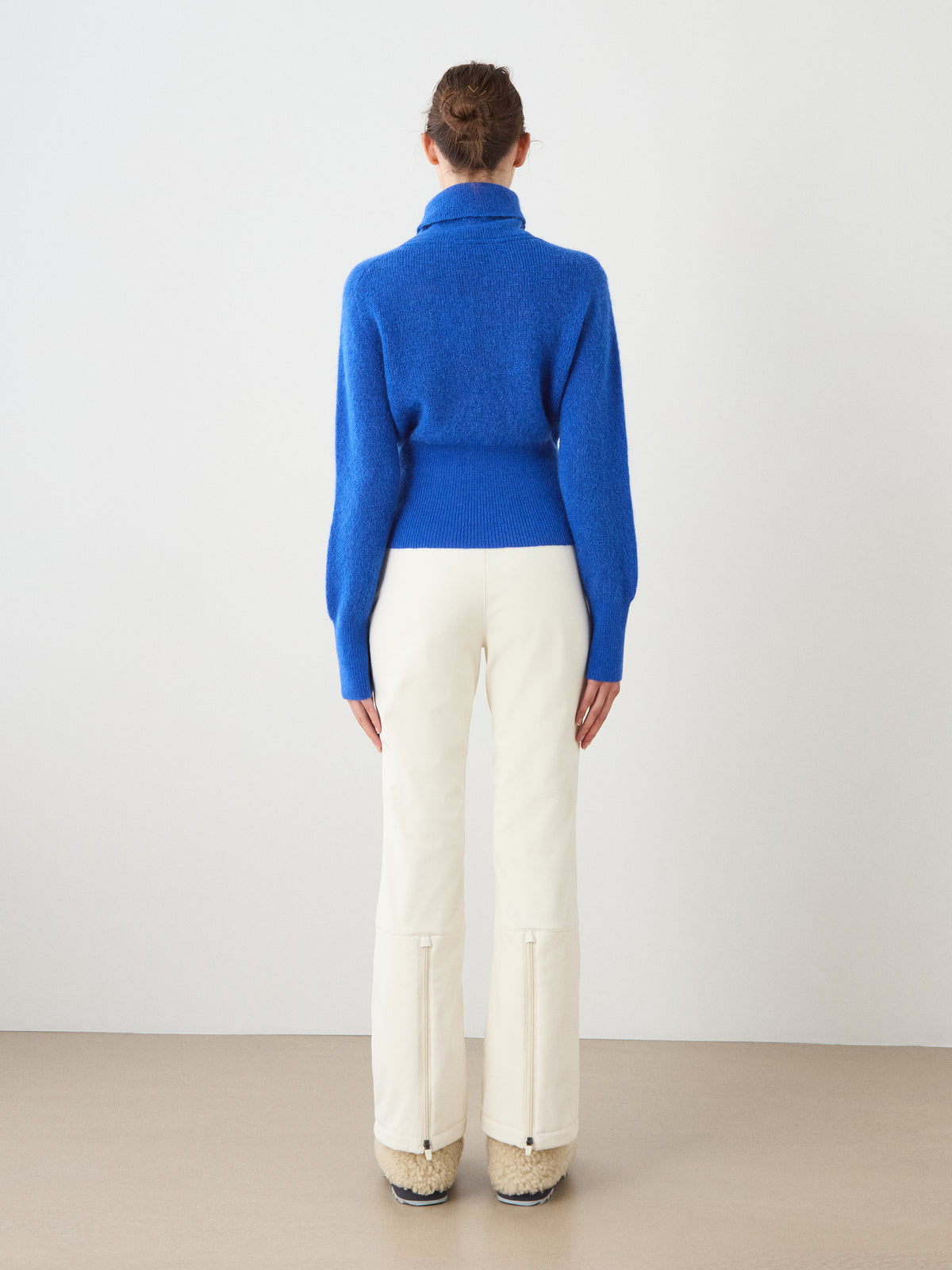 A person stands with their back to the camera, wearing the We Norwegians Pudder Sweater Women—a bright blue long-sleeve cropped turtleneck—paired with cream-colored pants and beige shoes, set against a plain light background.