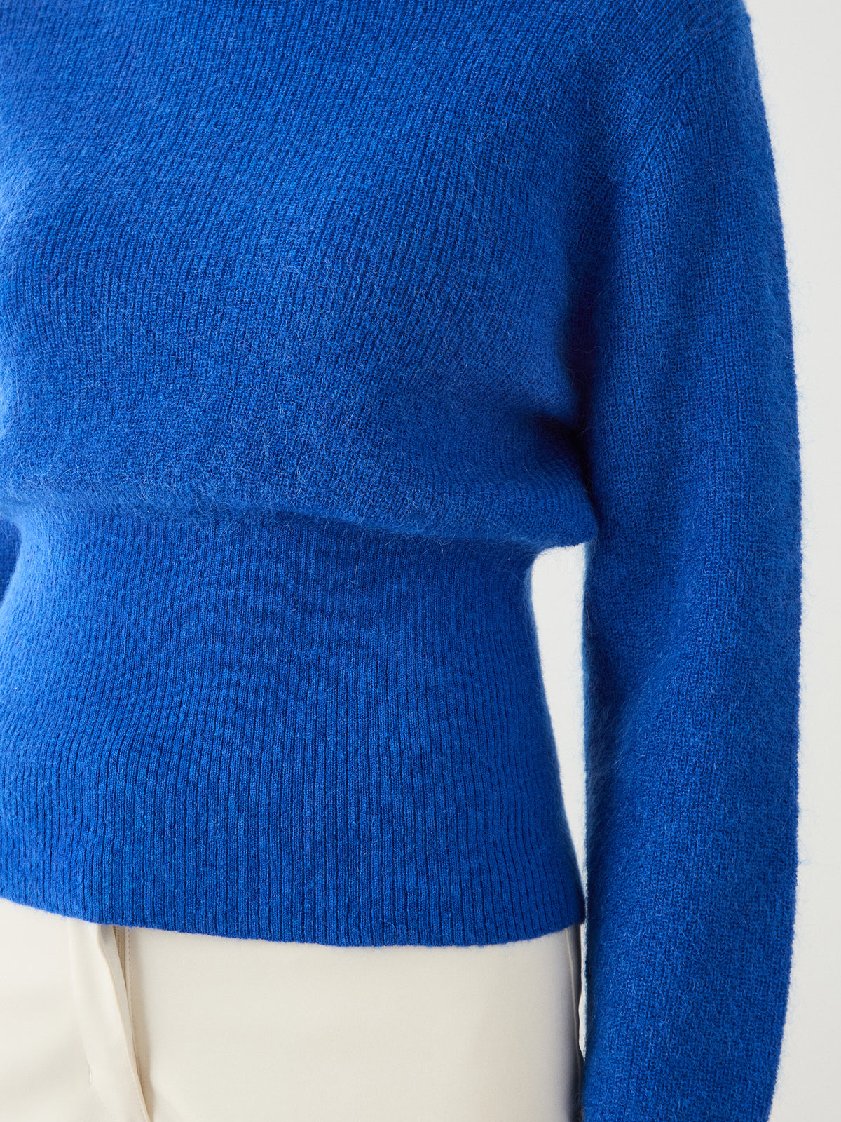 A person wearing the We Norwegians Pudder Sweater Women—a bright blue, long-sleeved, knit cropped sweater crafted from mohair yarn—paired with cream-colored pants, shown from the shoulders to the hips.