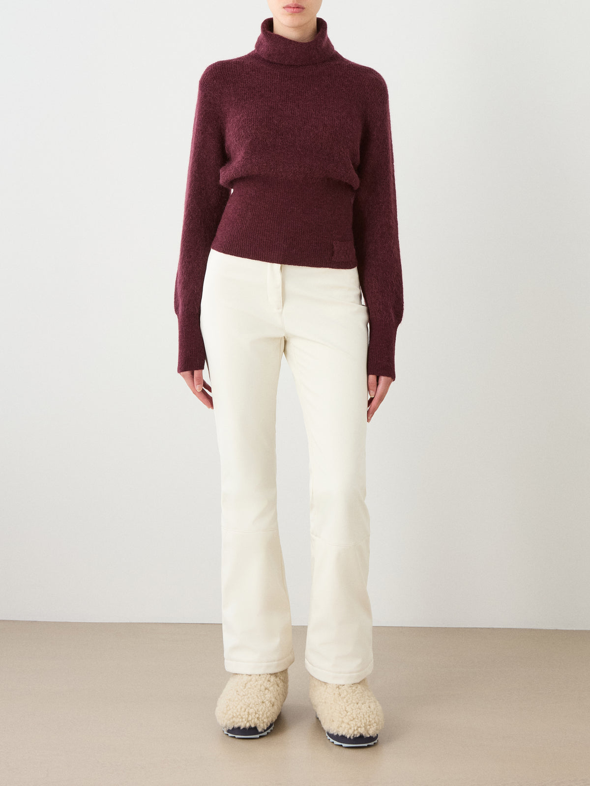 A person, head not visible, stands against a plain background wearing the We Norwegians Pudder Sweater Women in burgundy mohair yarn, paired with white pants and cream fuzzy slippers.