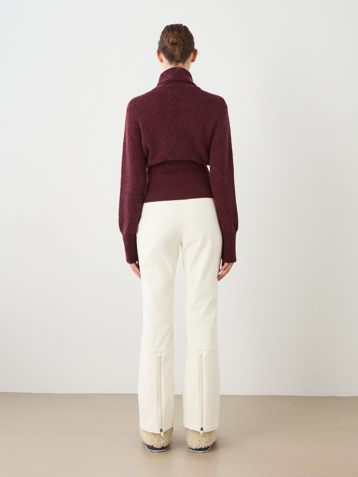 A woman stands facing away, wearing the We Norwegians Pudder Sweater Women—a maroon turtleneck crafted from soft mohair yarn—paired with white pants and beige fur-lined shoes, against a plain light background.