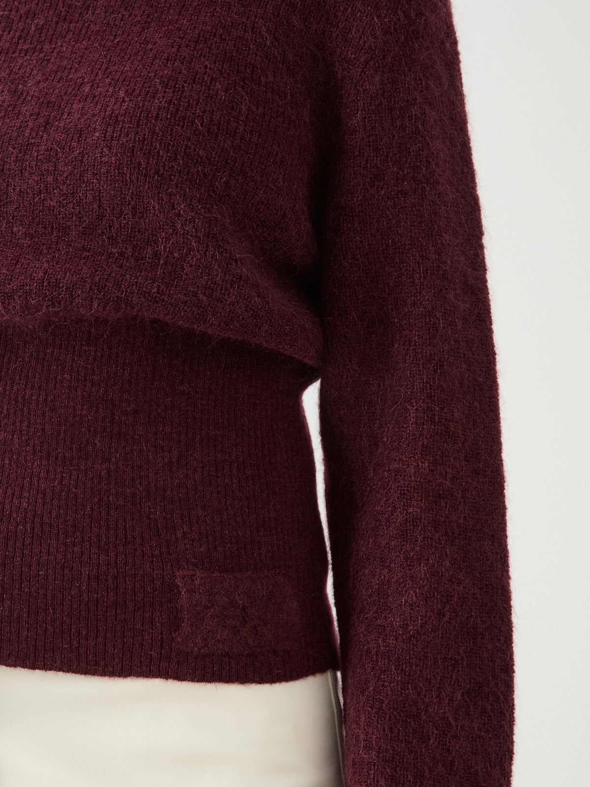 A close-up of a person wearing the We Norwegians Pudder Sweater Women, a dark maroon fuzzy cropped sweater made from soft mohair yarn, paired with light-colored pants, showing the upper body from shoulder to waist against a plain background.