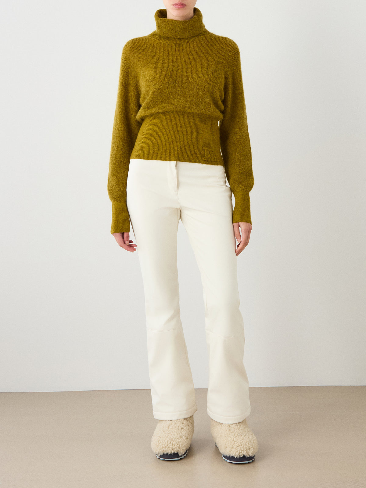 A person wearing the We Norwegians Pudder Sweater Women in mustard, crafted from cozy mohair yarn, paired with white pants and cream-colored fuzzy slippers, stands against a plain light background. The person’s head is not visible.