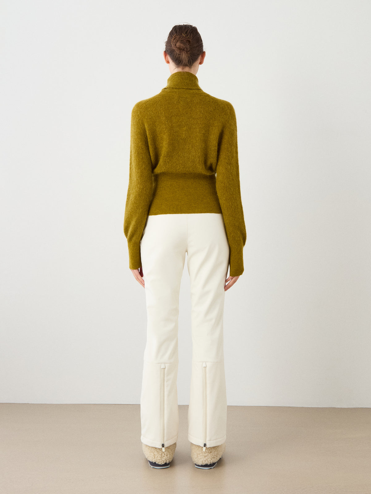 Facing away from the camera, a person wears the We Norwegians Pudder Sweater Women in olive green—a mohair yarn turtleneck cropped sweater—paired with white ankle-zip pants and beige shoes, set against a neutral background.