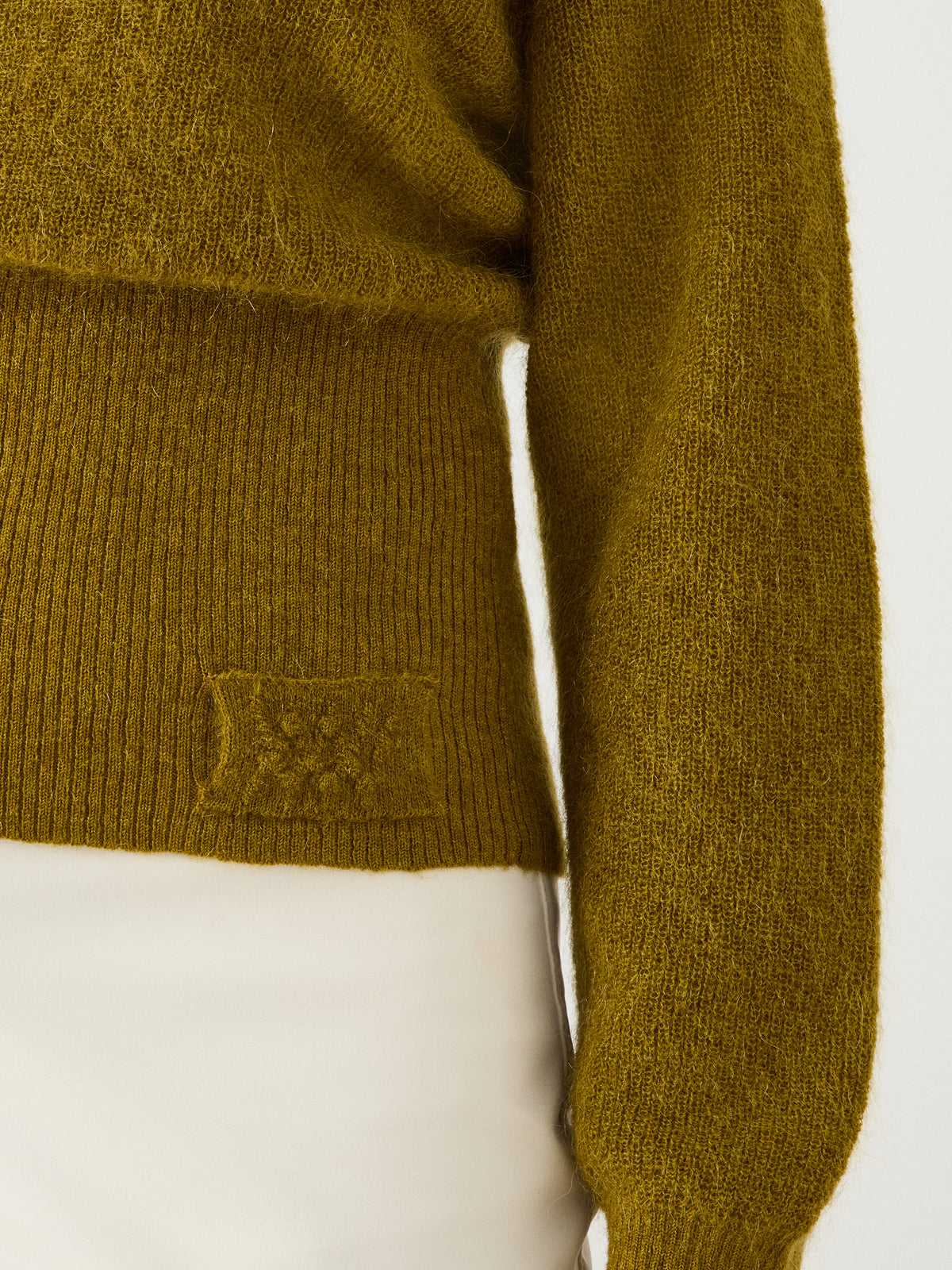 A close-up of a person wearing the We Norwegians Pudder Sweater Women in olive green—a mohair-knit design with a ribbed waistband and a small textured patch—paired with light-colored pants. Only part of the torso and one arm are visible.