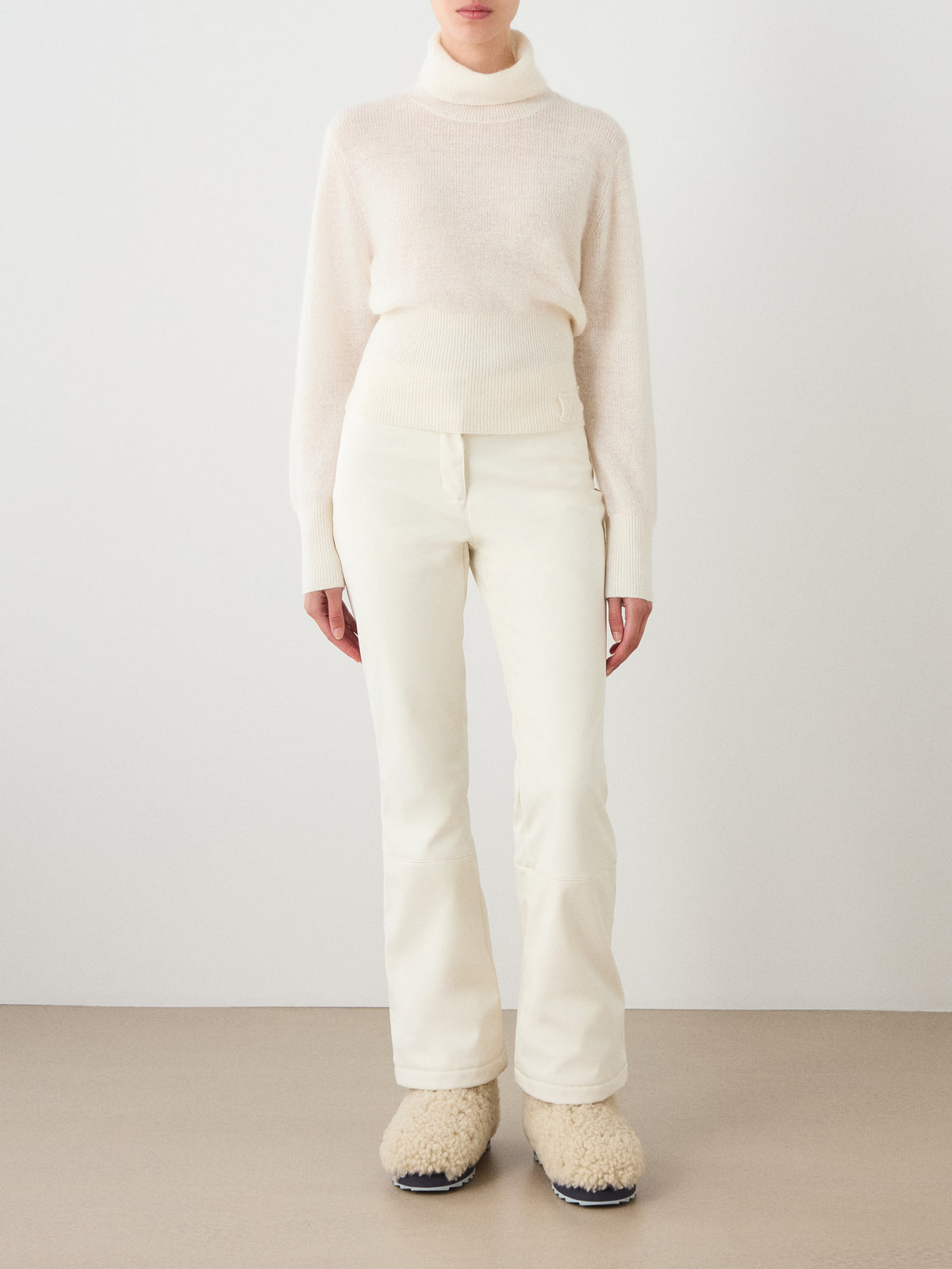 A person stands against a plain background wearing the We Norwegians Pudder Sweater Women, an off-white turtleneck knit from soft mohair yarn. They pair it with matching cream pants and fuzzy cream slippers with black soles. Their face is not visible.