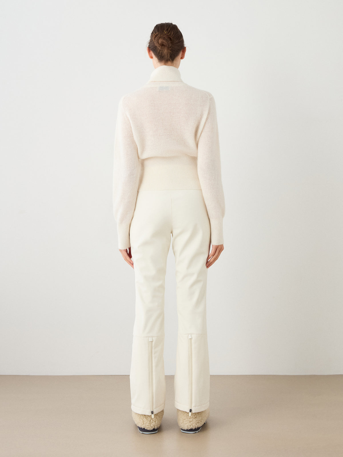 A person stands facing away against a plain light background, wearing the We Norwegians Pudder Sweater Women—a light cream cropped sweater crafted from mohair yarn—paired with high-waisted white pants and cream boots with textured soles.