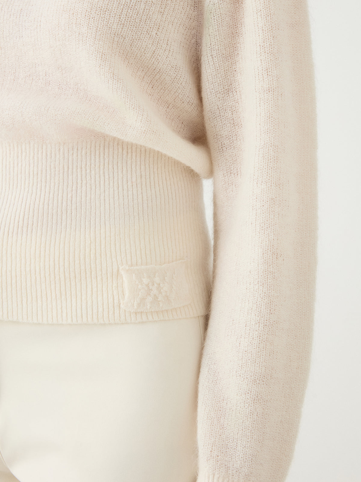 Close-up of a person wearing the cream-colored We Norwegians Pudder Sweater Women, a knit cropped sweater with ribbed hem crafted from soft mohair yarn, paired with off-white pants. Only the torso and one arm are visible against a plain light background.