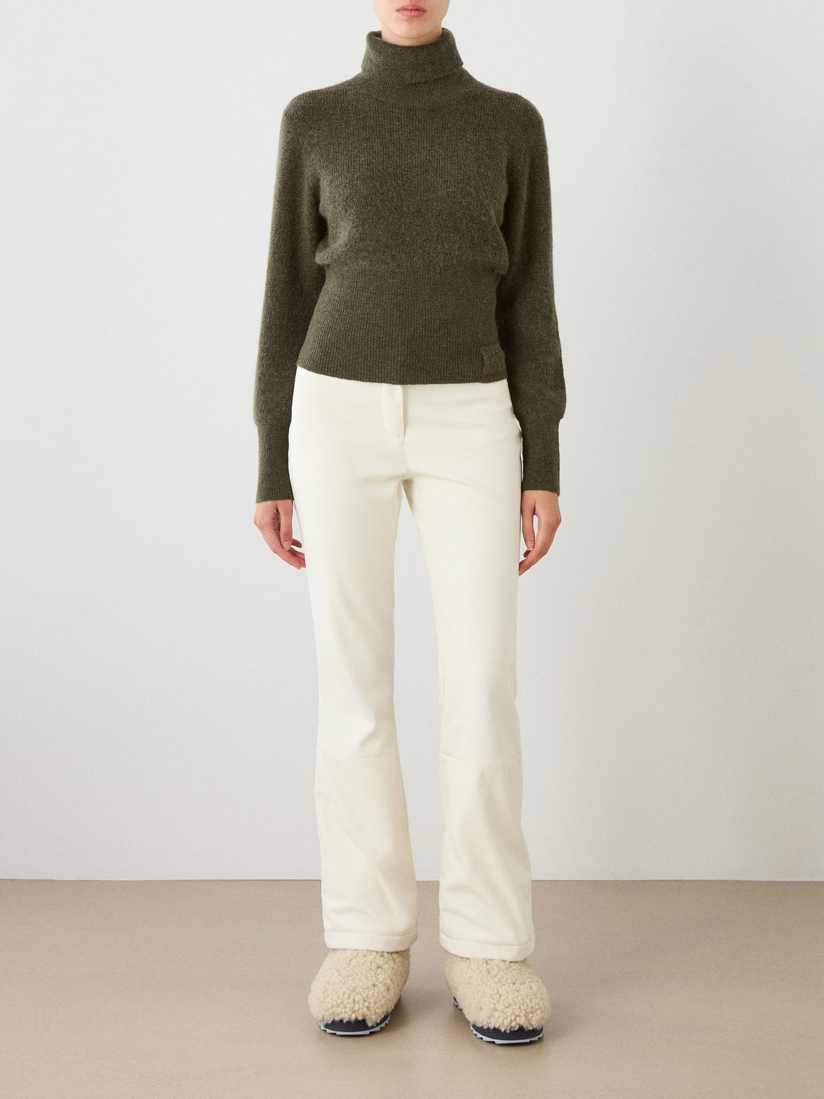 A person wearing the We Norwegians Pudder Sweater Women in dark green, paired with white pants and fuzzy beige slip-on shoes, stands against a plain wall, facing forward.