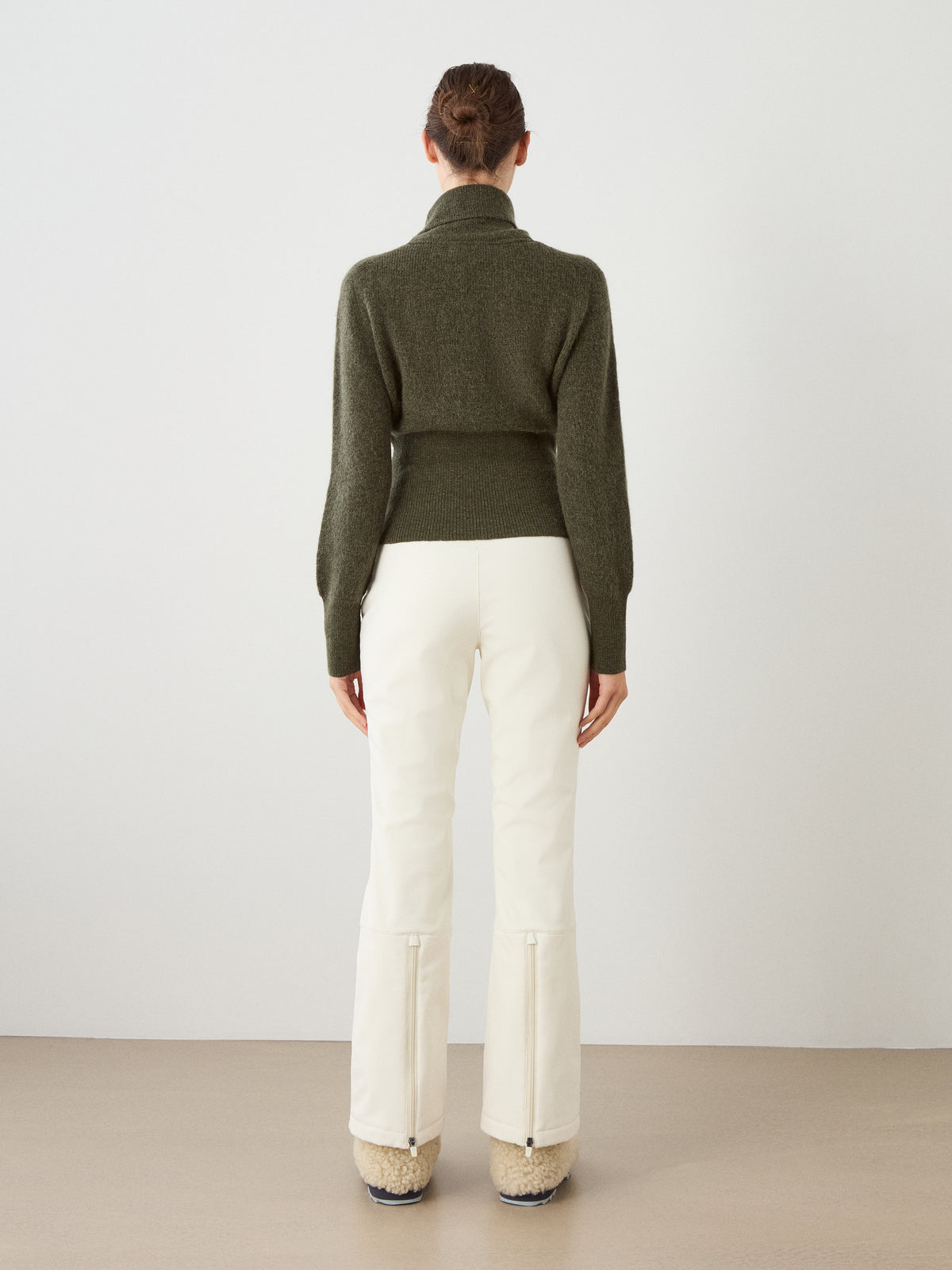 A person stands facing away from the camera, wearing the We Norwegians Pudder Sweater Women—a dark green cropped sweater crafted from mohair yarn—paired with white pants featuring back zippers and beige fuzzy shoes, against a plain background.