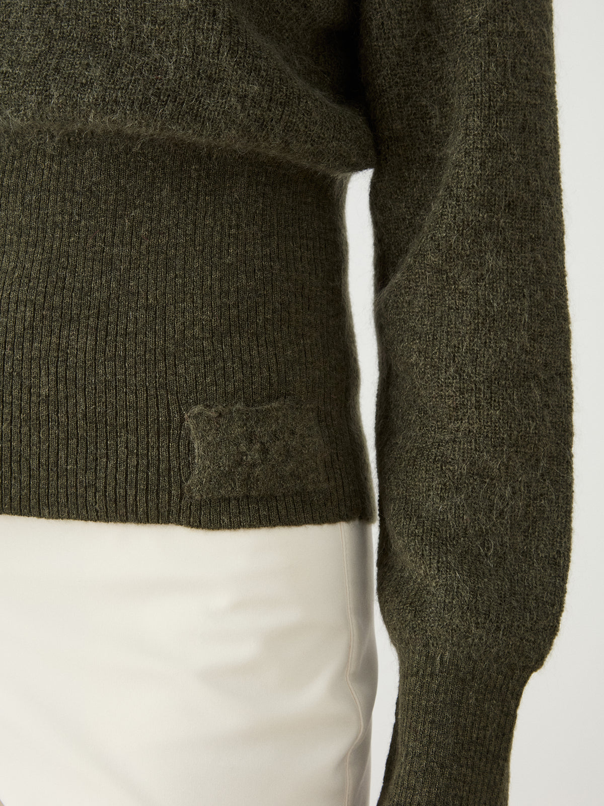 A close-up of a person wearing the We Norwegians Pudder Sweater Women in olive green, featuring a turtleneck, ribbed hem, and long sleeves, paired with white pants. Only the torso and right arm are visible.
