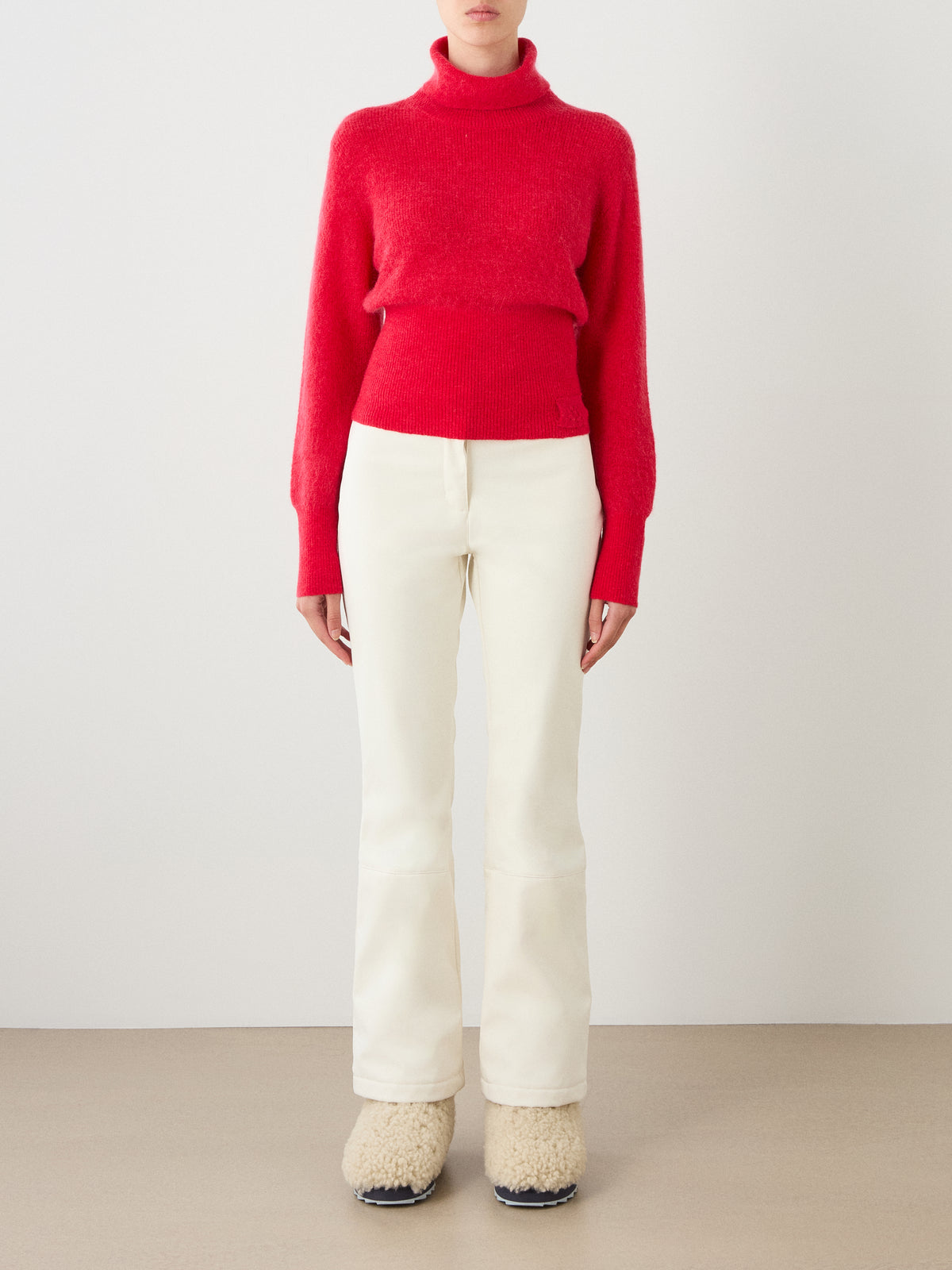 A person stands against a plain wall wearing the We Norwegians Pudder Sweater Women—a red mohair yarn turtleneck—paired with off-white pants and fuzzy cream slippers. Their hands are at their sides and their face is not visible.