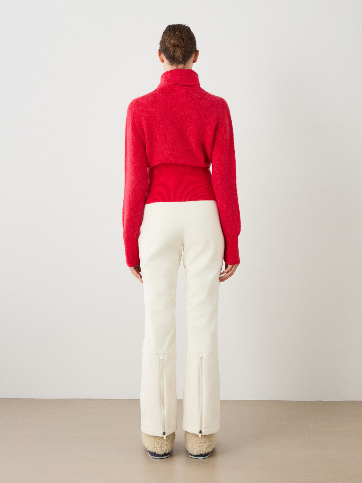 A person stands facing away, wearing the We Norwegians Pudder Sweater Women—a red turtleneck cropped sweater knit from mohair yarn—paired with white pants featuring back zippers and beige fuzzy boots, set against a plain light background.