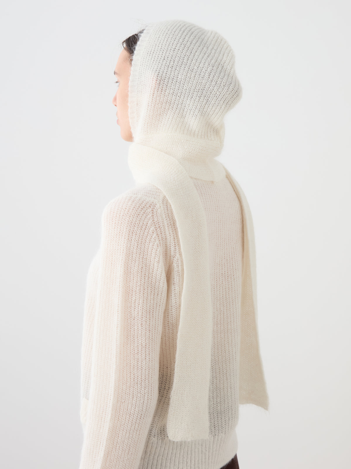 A person wearing a cosy, cream-colored We Norwegians Pudder Balaclava with long scarf ends draped over a matching sweater.