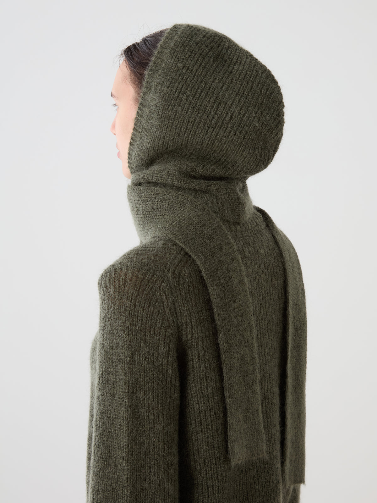 A person wearing a dark olive green Pudder Balaclava by We Norwegians, featuring a scarf-like extension hanging down the back as a cosy winter accessory.