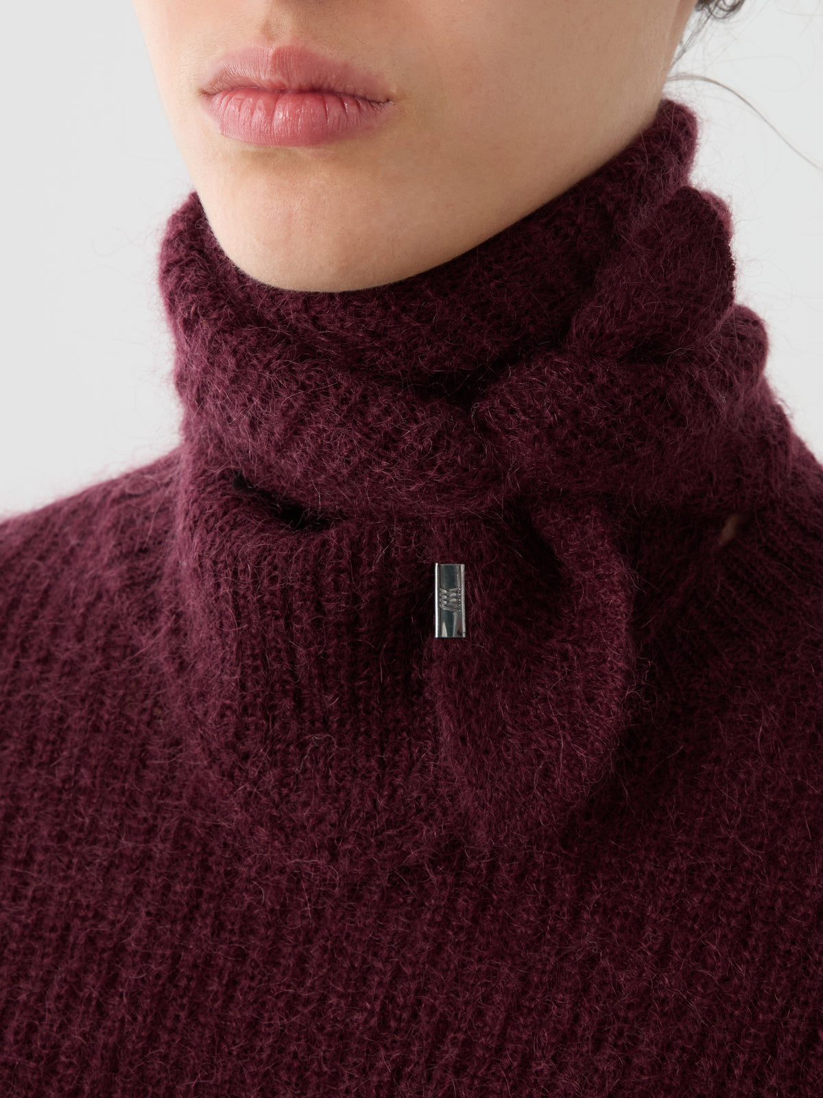 A close-up of a person wearing a textured, deep burgundy turtleneck sweater with a small metallic tag near the collar, styled with the luxurious Pudder Scarf by We Norwegians for an added touch of designer winter accessory elegance.