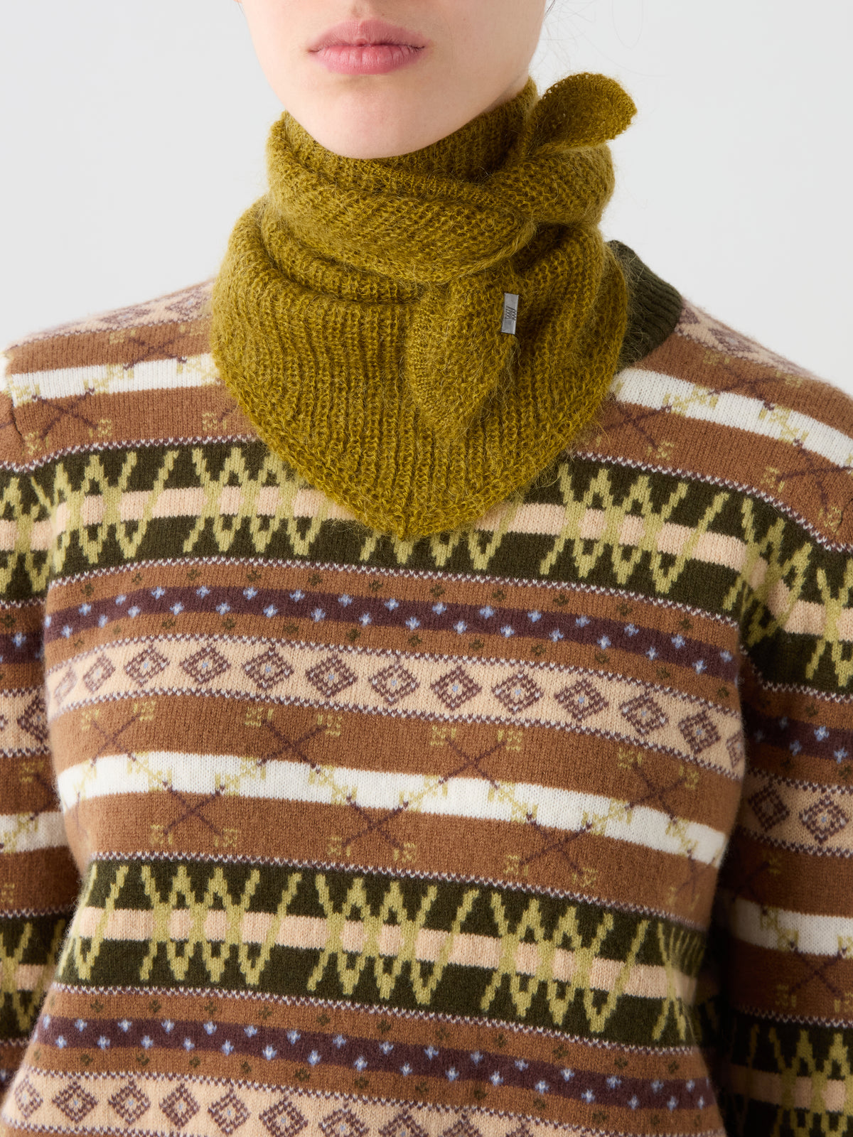 A person wearing a patterned brown, green, and beige sweater and a textured olive green We Norwegians Pudder Scarf—crafted from soft mohair—tied around their neck. Their face is partially visible from the nose down.