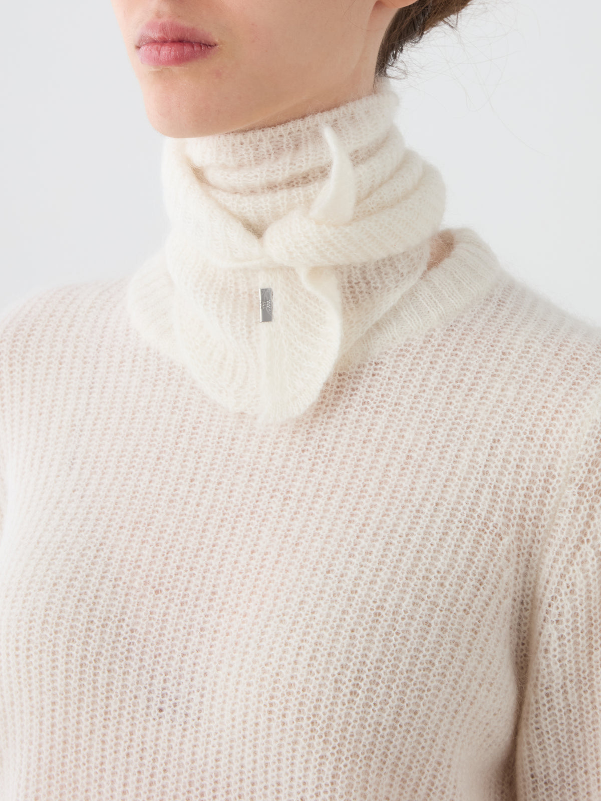 A person wearing a soft, white, textured knit sweater and the We Norwegians Pudder Scarf—a signature high-neck mohair scarf in a matching shade featuring a subtle branded tag—against a plain background; the perfect designer winter accessory.