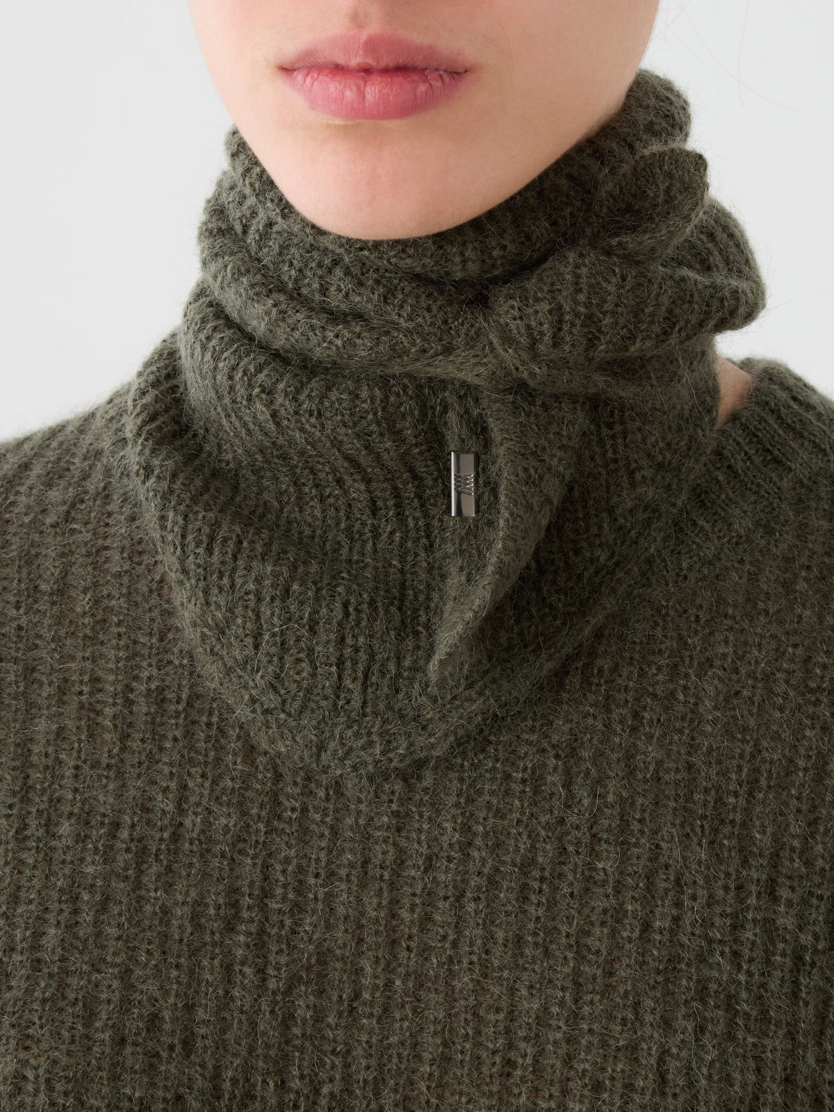 A person is shown wearing a textured olive green knit sweater, accessorized with the We Norwegians Pudder Scarf wrapped around their neck and secured with a small metal pin. Only the lower half of their face is visible.