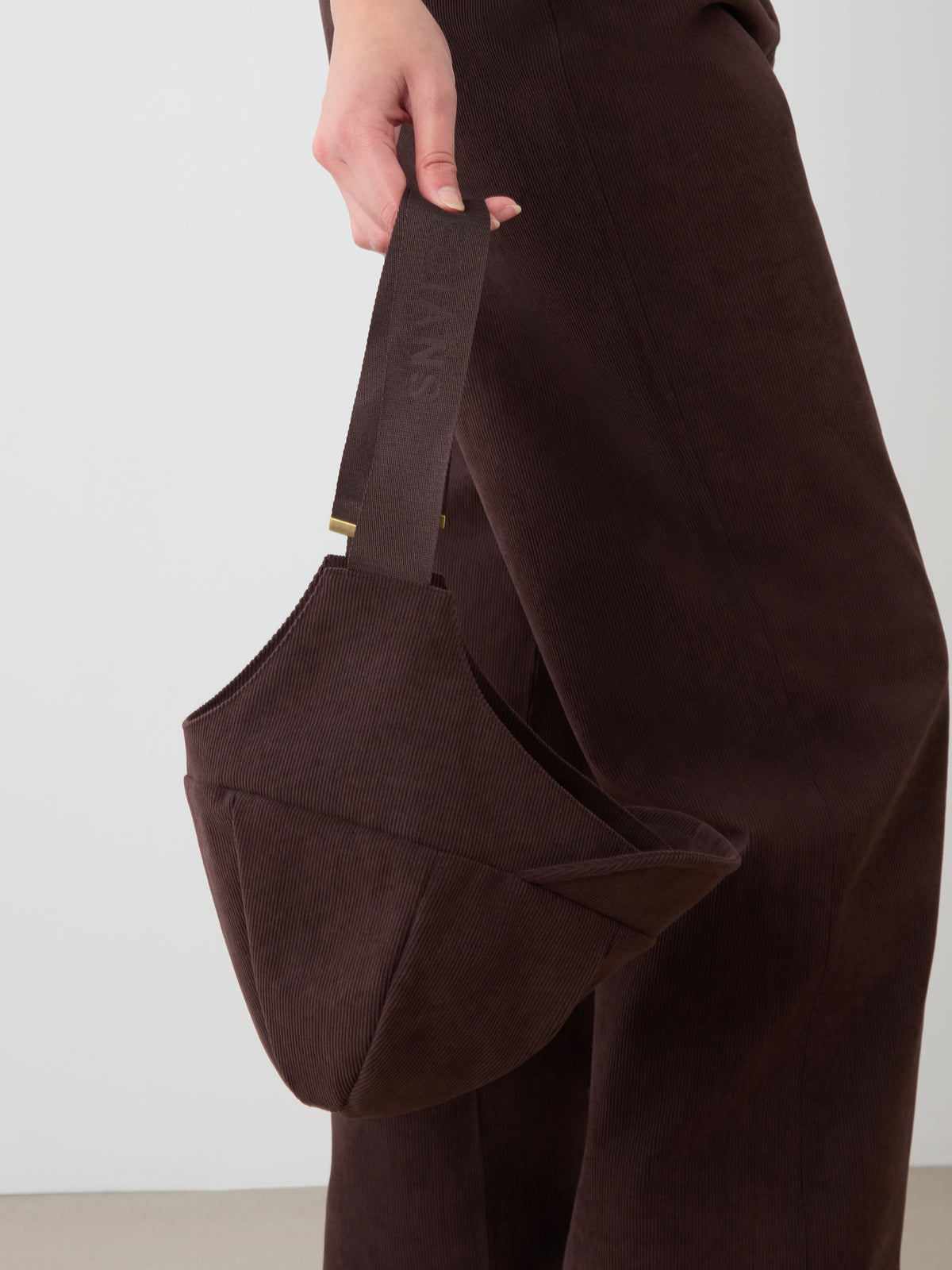 A person in dark brown baby corduroy pants holds the uniquely shaped Britt Bonnet by We Norwegians, gripping its short handle against a neutral background.