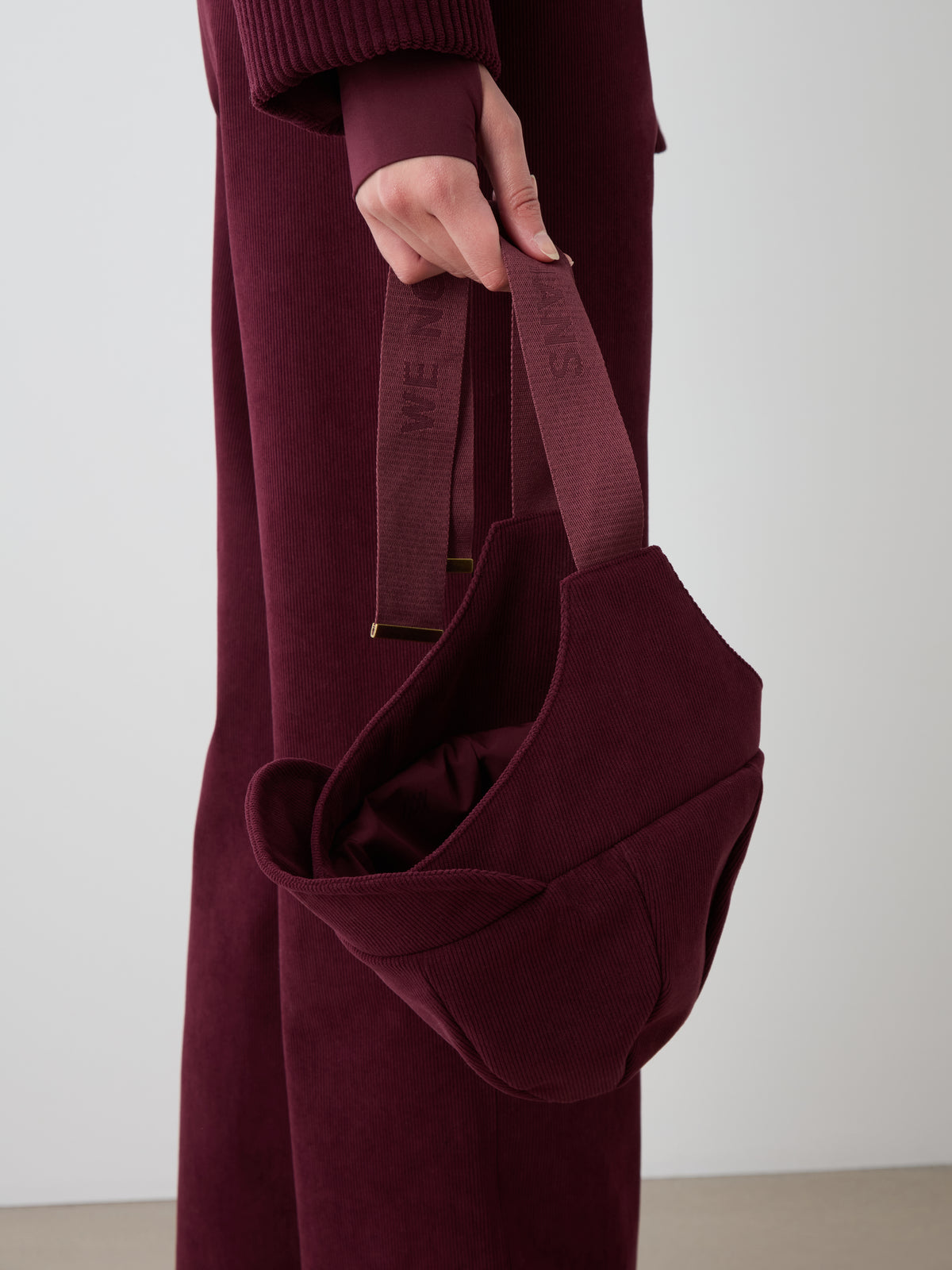 A person in a burgundy baby corduroy outfit holds the Britt Bonnet by We Norwegians in their left hand against a plain, neutral background—perfect for highlighting winter alpine accessories.
