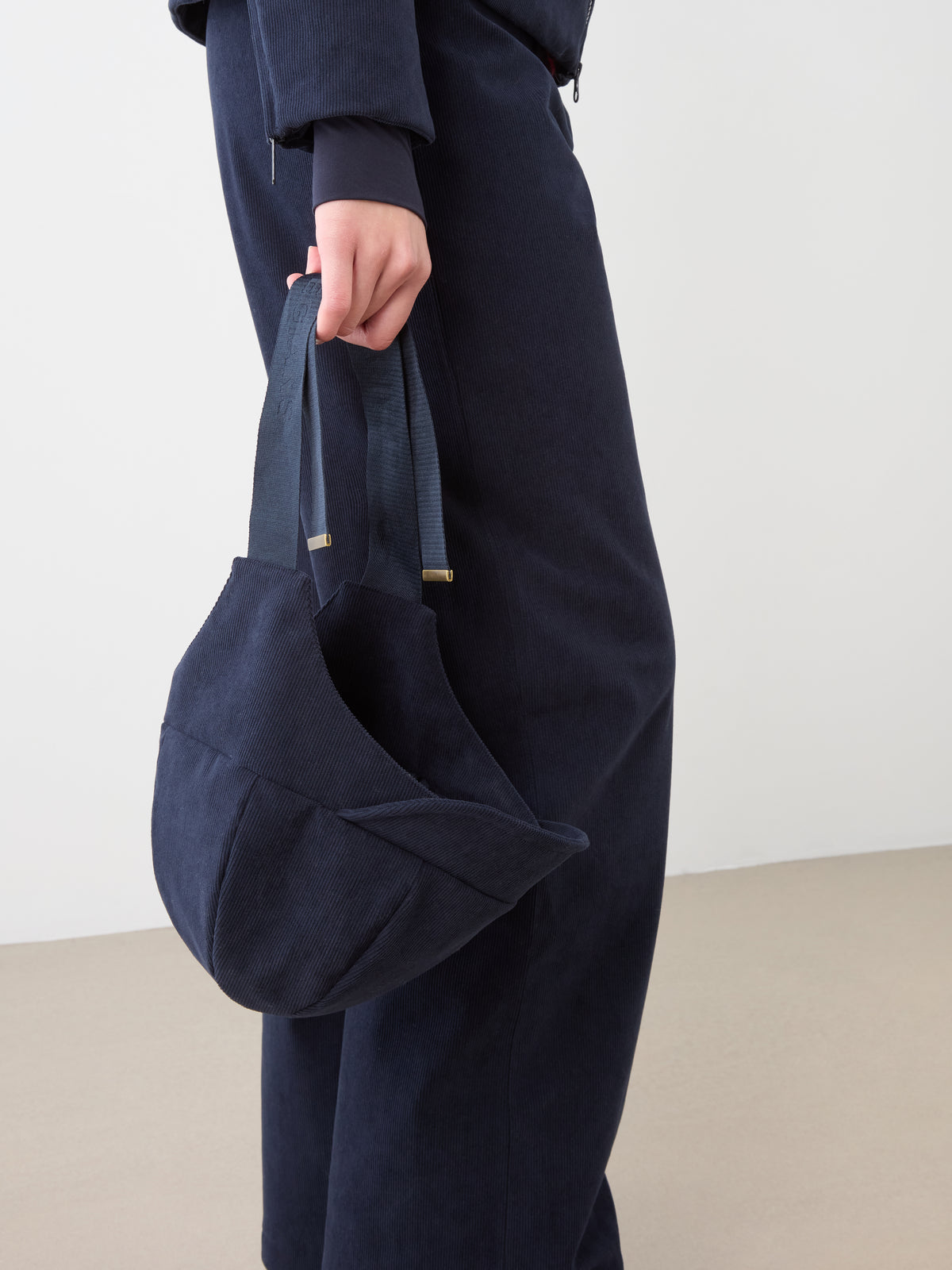 A person wears a navy blue Britt Bonnet by We Norwegians and holds a matching fabric handbag with short handles, standing indoors against a plain background.