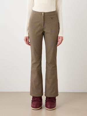 Wearing Glenshee Ski Pant Women by We Norwegians in brown and beige houndstooth, paired with a white long-sleeve top and maroon boots; shown from shoulders down against a neutral background.