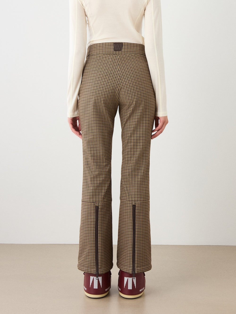 Person stands facing away, wearing a cream long-sleeve top, We Norwegians Glenshee Ski Pant Women in brown plaid with zippered hems, and burgundy boots with white accents, against a neutral background.