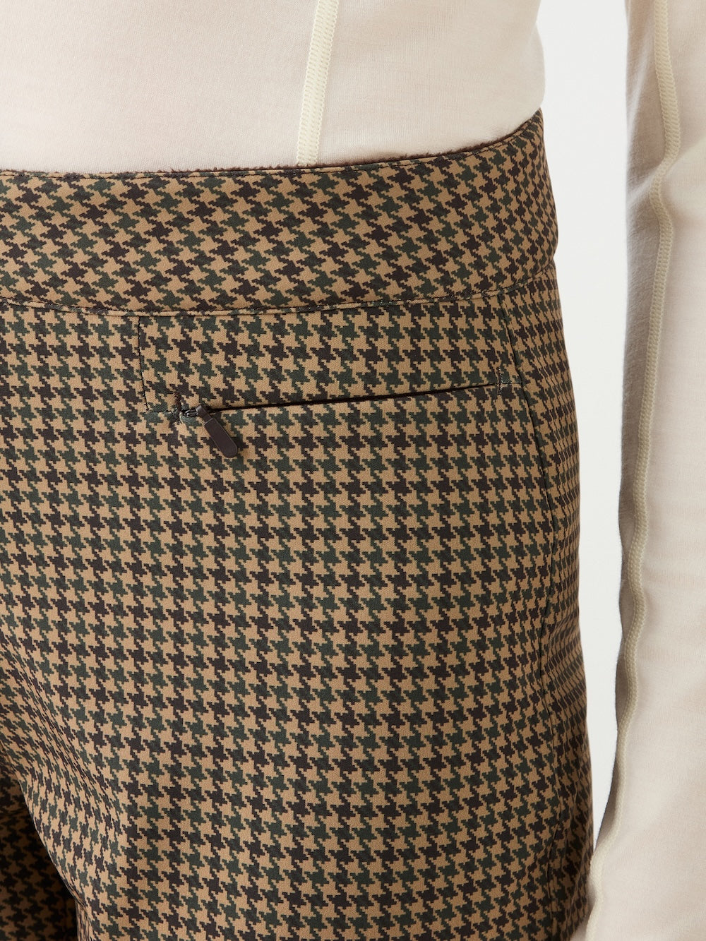 A close-up of a person wearing brown and black houndstooth Glenshee Ski Pant Women by We Norwegians with a cream long-sleeve top. Features a small welt pocket for added style.