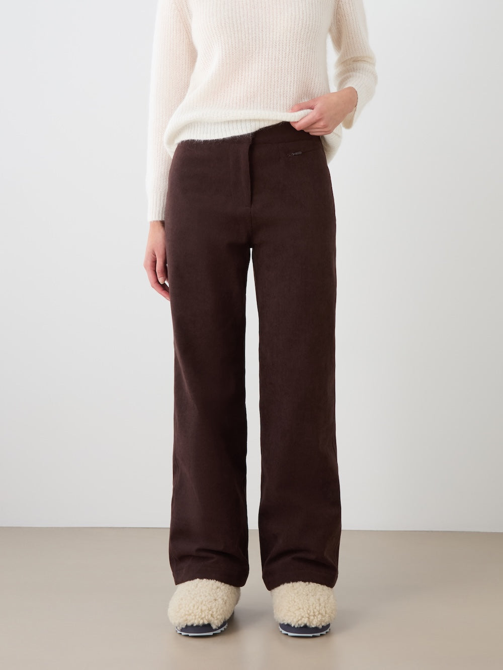 A person wearing a white sweater, the We Norwegians Britt Palazzo Ski Pant for women in dark brown, and beige fuzzy slippers stands against a plain light background.
