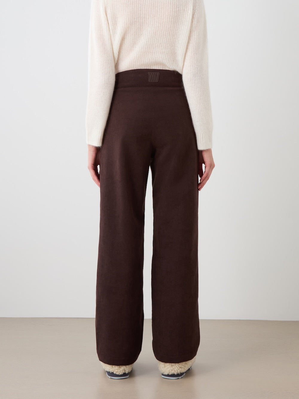 A person is standing facing away, wearing a cream long-sleeve sweater with We Norwegians Britt Palazzo Ski Pant Women in dark brown and fluffy slippers, against a plain light background.