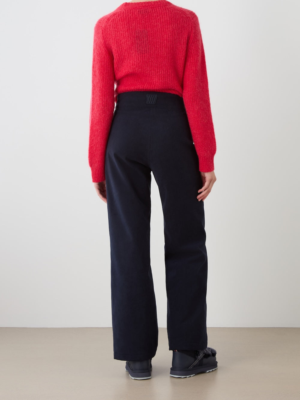 A person stands facing away from the camera, wearing a bright red long-sleeve sweater, We Norwegians Britt Palazzo Ski Pant for women, and black shoes against a plain light background.