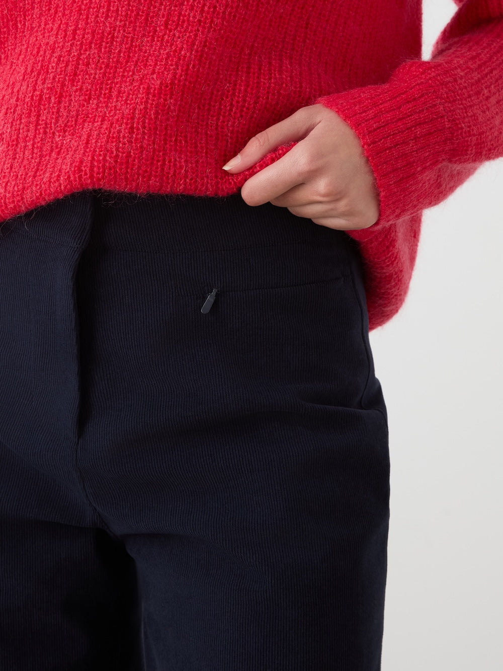 A woman is wearing the We Norwegians Britt Palazzo Ski Pant, styled in a dark navy high-waisted cut, with one hand tucked casually into the pocket; the photo highlights the details of both the pants and her bright red sweater.