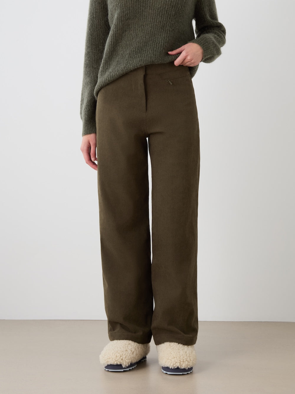 A person wearing a green knit sweater, the We Norwegians Britt Palazzo Ski Pant Women in a loose brown style, and cream-colored fuzzy slippers stands against a plain light background. Their face is not visible.