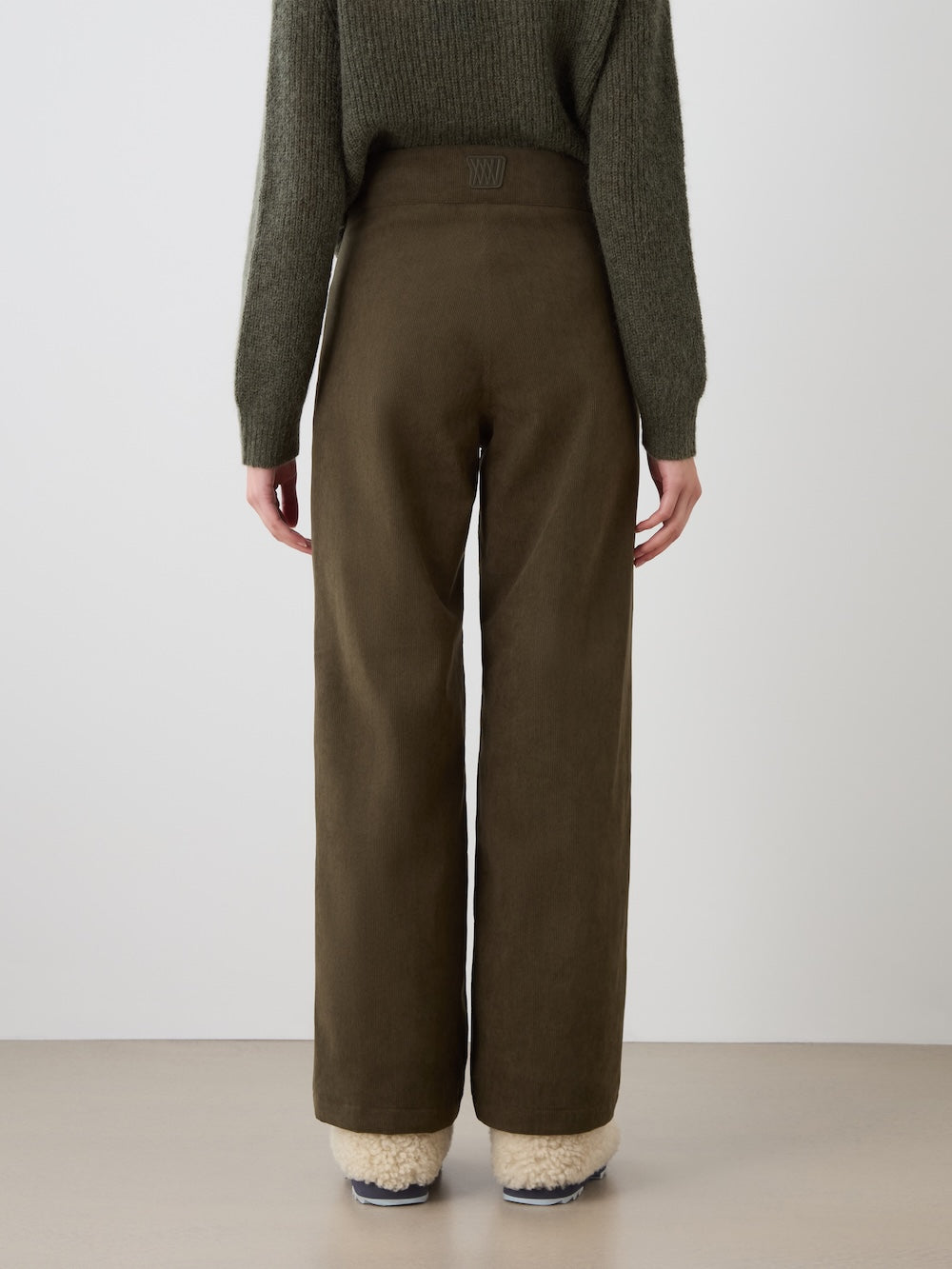 A person stands with their back to the camera, wearing a dark green sweater, We Norwegians Britt Palazzo Ski Pant Women in olive green, and beige fuzzy slippers, set against a plain light background.