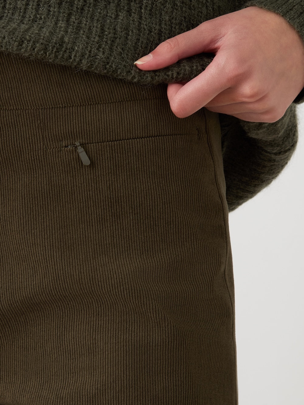 A close-up of a person wearing the We Norwegians Britt Palazzo Ski Pant Women in olive green baby corduroy and a textured green sweater, with one hand gently tugging at the sweater hem near a zippered pocket.