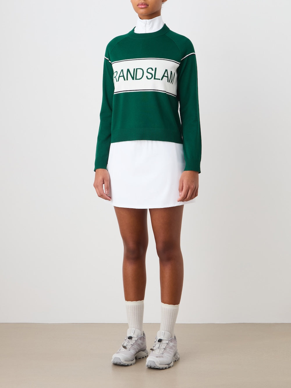 A person wearing a white tennis dress and the We Norwegians Grand Slam Sweater Women, paired with white socks and gray sneakers, stands against a plain light background. The image does not show their head.