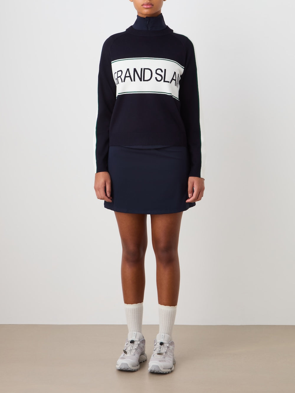Wearing the We Norwegians Grand Slam Sweater Women in navy with a matching skirt, white socks, and gray sneakers creates a vintage athletic look—ideal as an après sport layer against a simple backdrop.