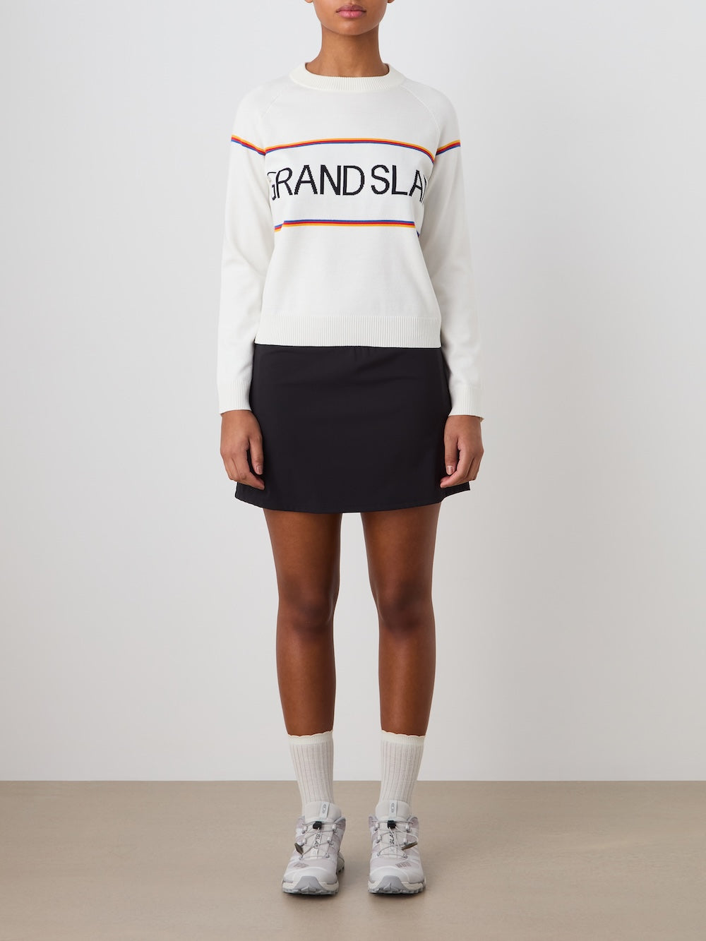 A woman stands against a plain background wearing the We Norwegians Grand Slam Sweater, paired with a short black skirt, white socks, and gray athletic sneakers. Her hands are at her sides.