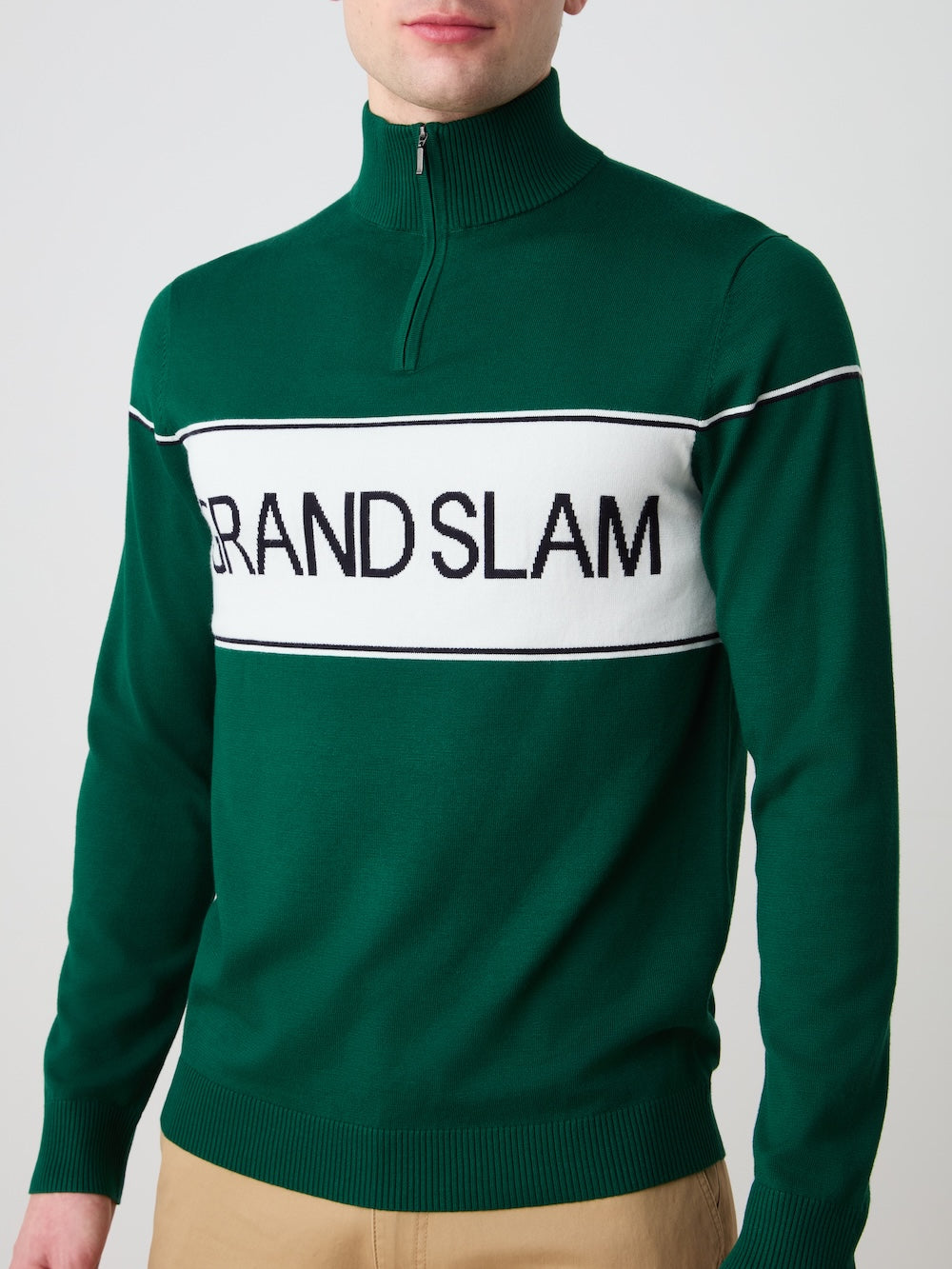 A man wears the We Norwegians Grand Slam Sweater Men, a green quarter-zip with a white stripe and bold "GRAND SLAM" lettering, paired with tan pants against a plain light gray background.