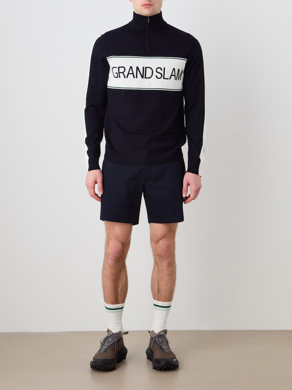 A person stands against a plain background, wearing the We Norwegians Grand Slam Sweater Men—a stylish après sport layer—paired with black shorts, white socks with green stripes, and black sneakers.