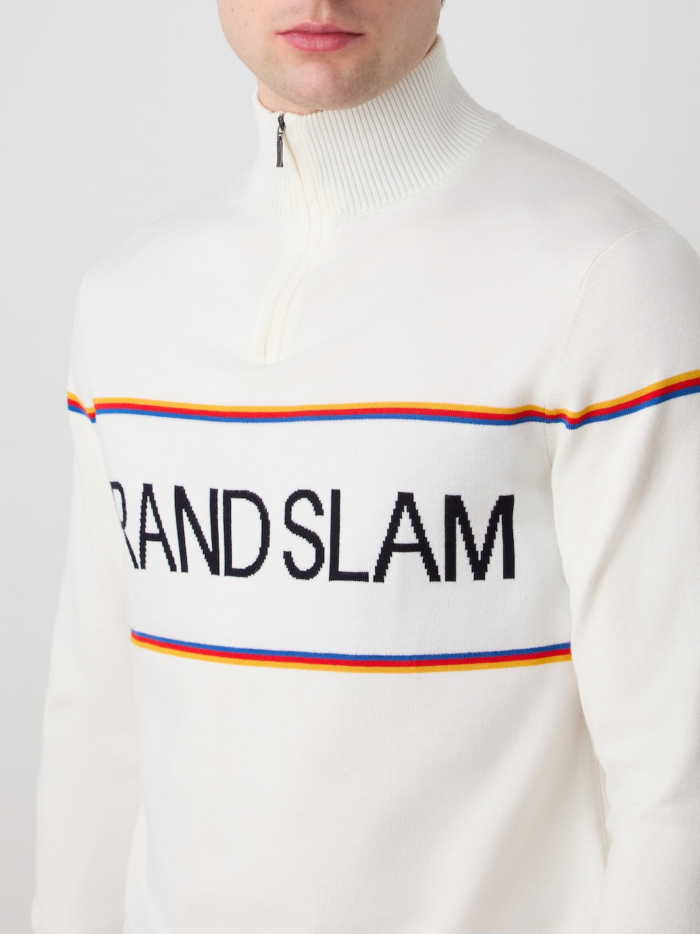 A person wears the We Norwegians Grand Slam Sweater Men in white, featuring "GRAND SLAM" in bold black letters on the chest, bordered by thin red, yellow, and blue stripes—a classic vintage athletic look. The face is partially visible.