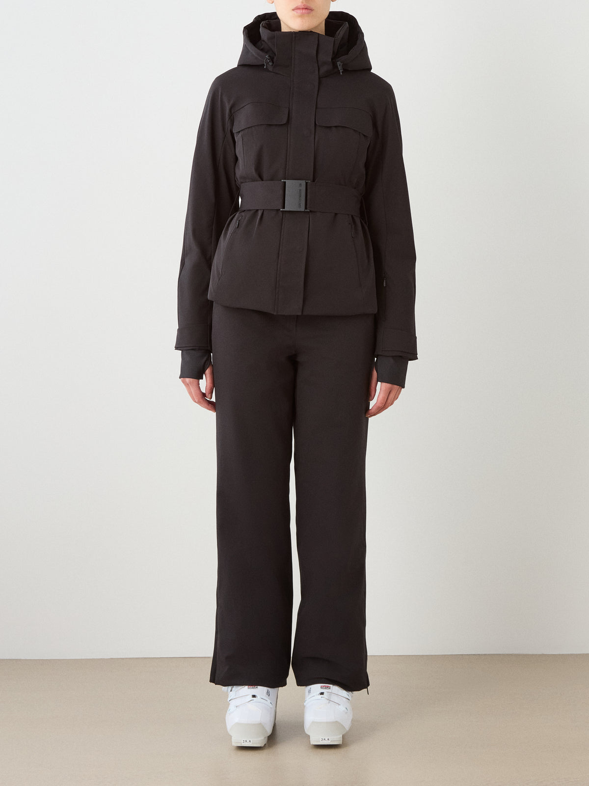 A person wearing a black We Norwegians Finse Ski Jacket Women and matching black pants faces forward, with white boots visible against a plain, light-colored background.
