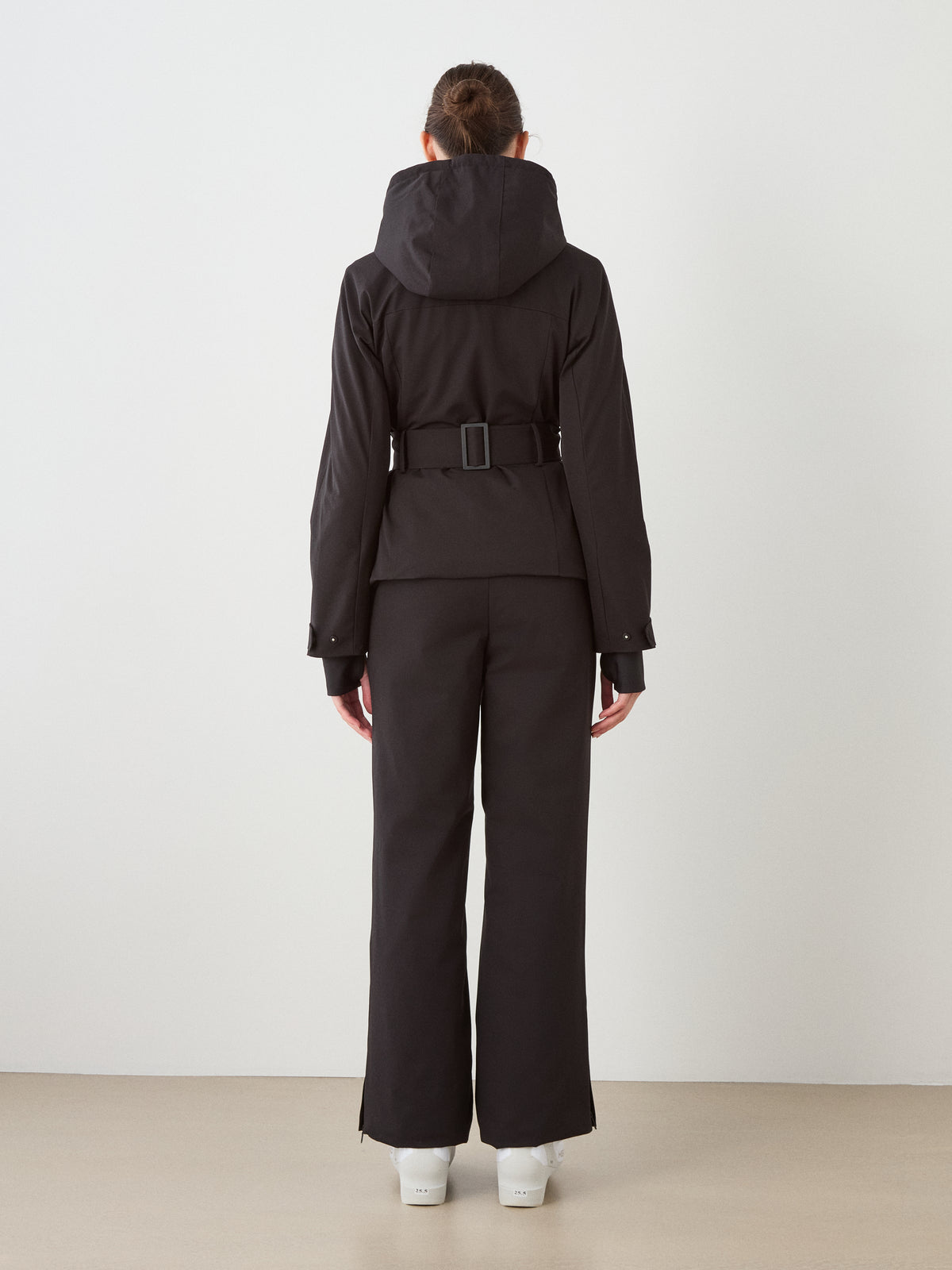 A person stands facing a white wall, wearing the black We Norwegians Finse Ski Jacket Women with hood up, matching black pants, and white boots—a sleek ski outfit with arms relaxed at their sides.