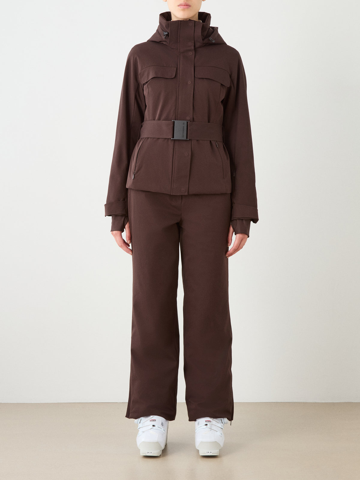 A person wears the We Norwegians Finse Ski Jacket Women in brown with matching pants and white boots, standing against a plain light background. Their face is not visible.
