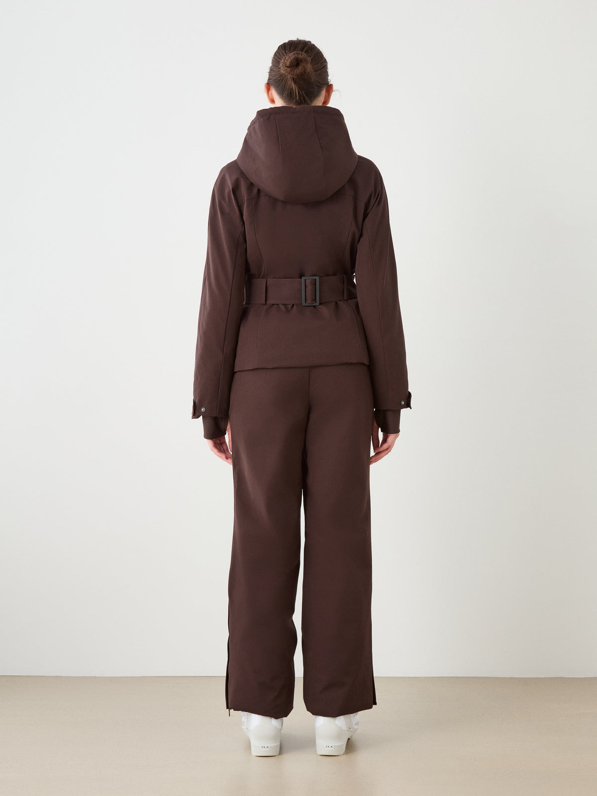 A person faces away, wearing the dark brown Finse Ski Jacket Women by We Norwegians with matching pants and white boots, creating an elegant look in a minimal, light-toned room.