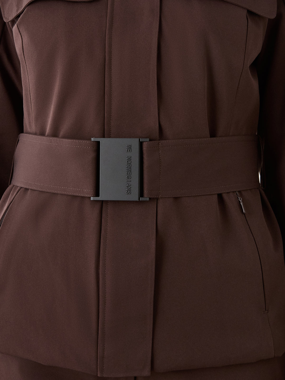 Close-up of a brown We Norwegians Finse Ski Jacket Women with a matching belt and rectangular black buckle featuring small text. This modern jacket includes zippered front pockets.
