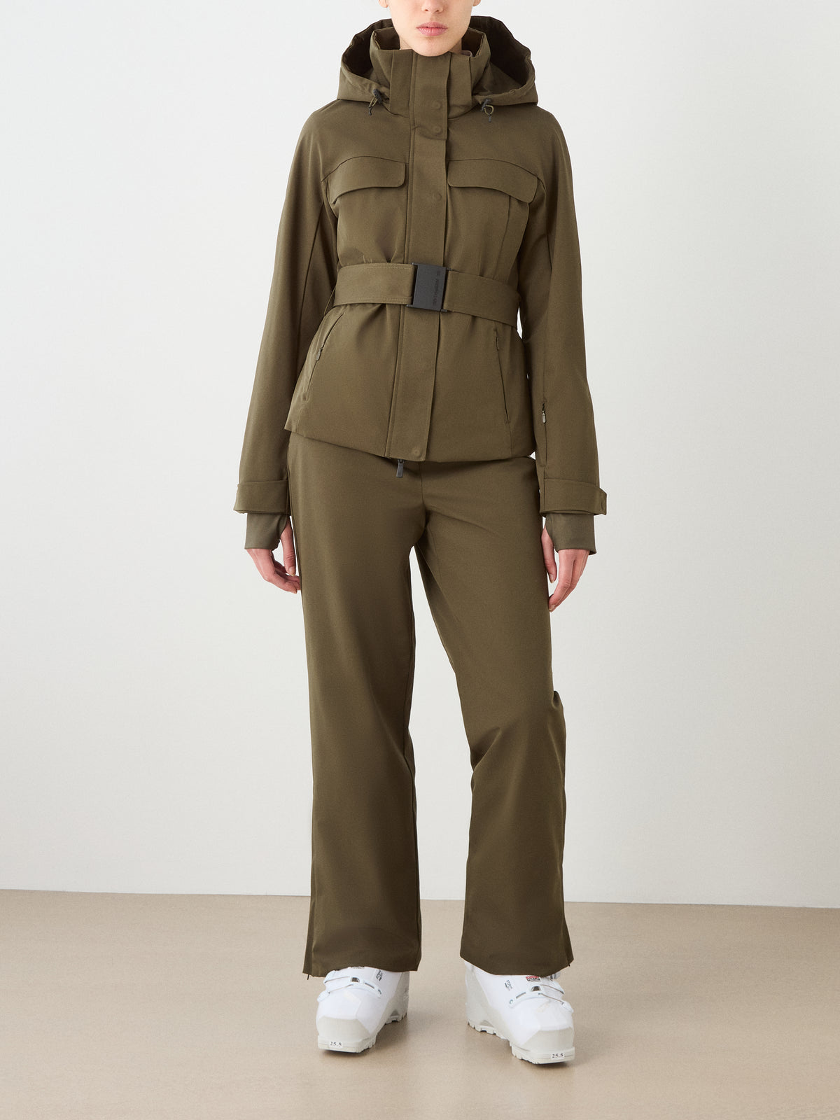 A person wearing the We Norwegians Finse Ski Jacket Women in olive green with matching pants and white ski boots, standing indoors against a plain light background—a chic ski look.
