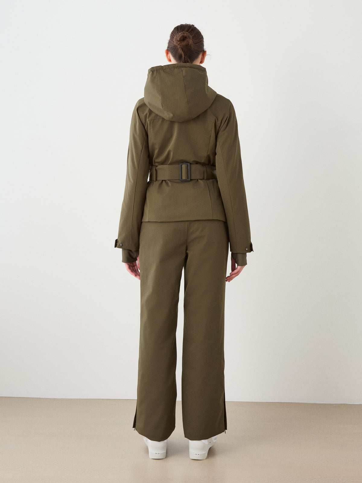 A person faces away, wearing the olive green Finse Ski Jacket Women by We Norwegians with a belt and matching pants, plus white boots, against a plain light wall and floor.