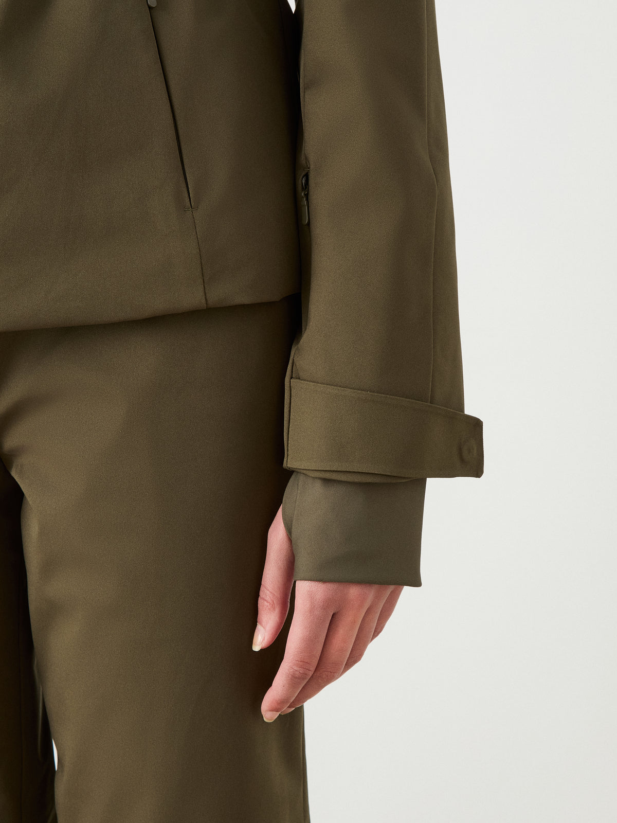 A person wears the Finse Ski Jacket Women by We Norwegians in olive green. The photo highlights a modern ski jacket’s buttoned strap on the sleeve, with a plain light background.